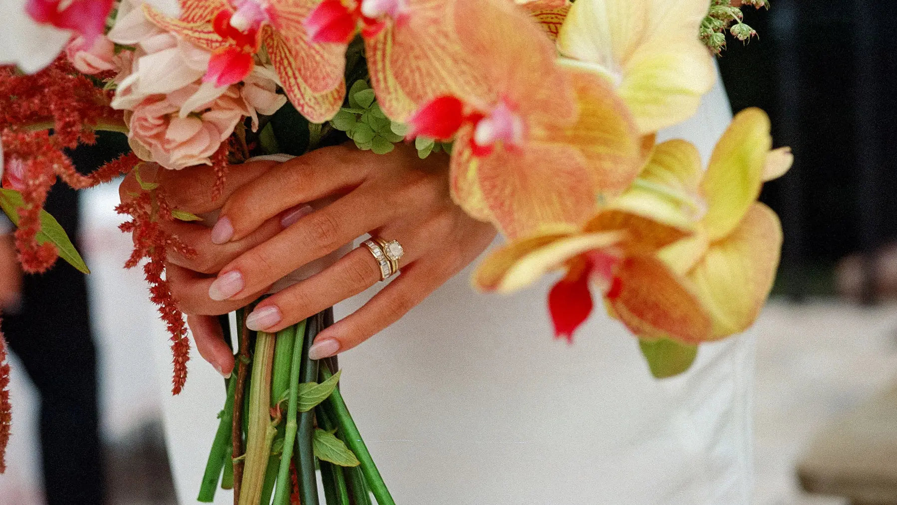 A North South Biggie Baguette Band stacked with a center stone ring with a cushion-cut white diamond on a woman's hand on her wedding day holding a bouquet