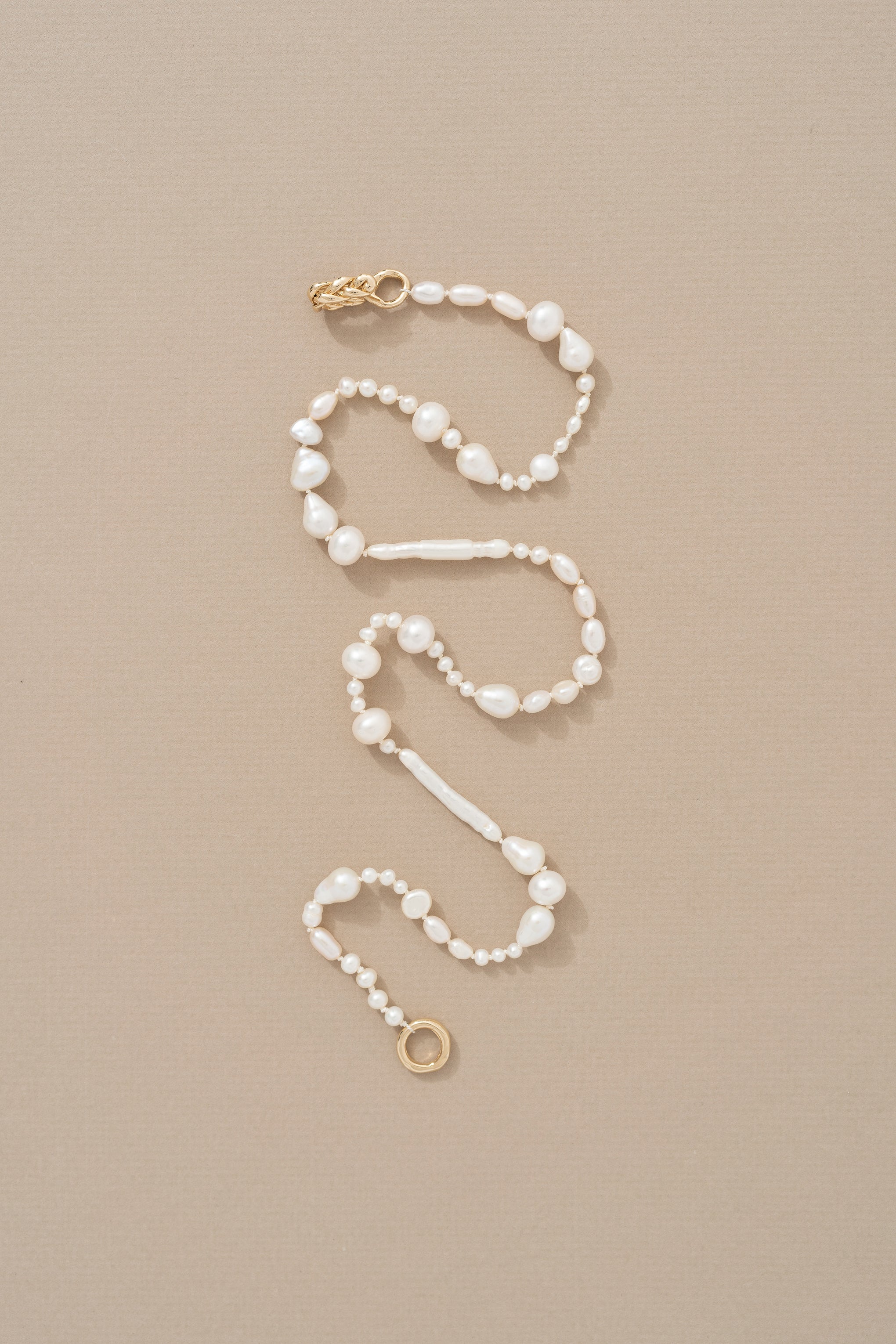 A knotted pearl necklace using a mix of different shaped pearls with a braided yellow gold closure, laid out on a neutral background.