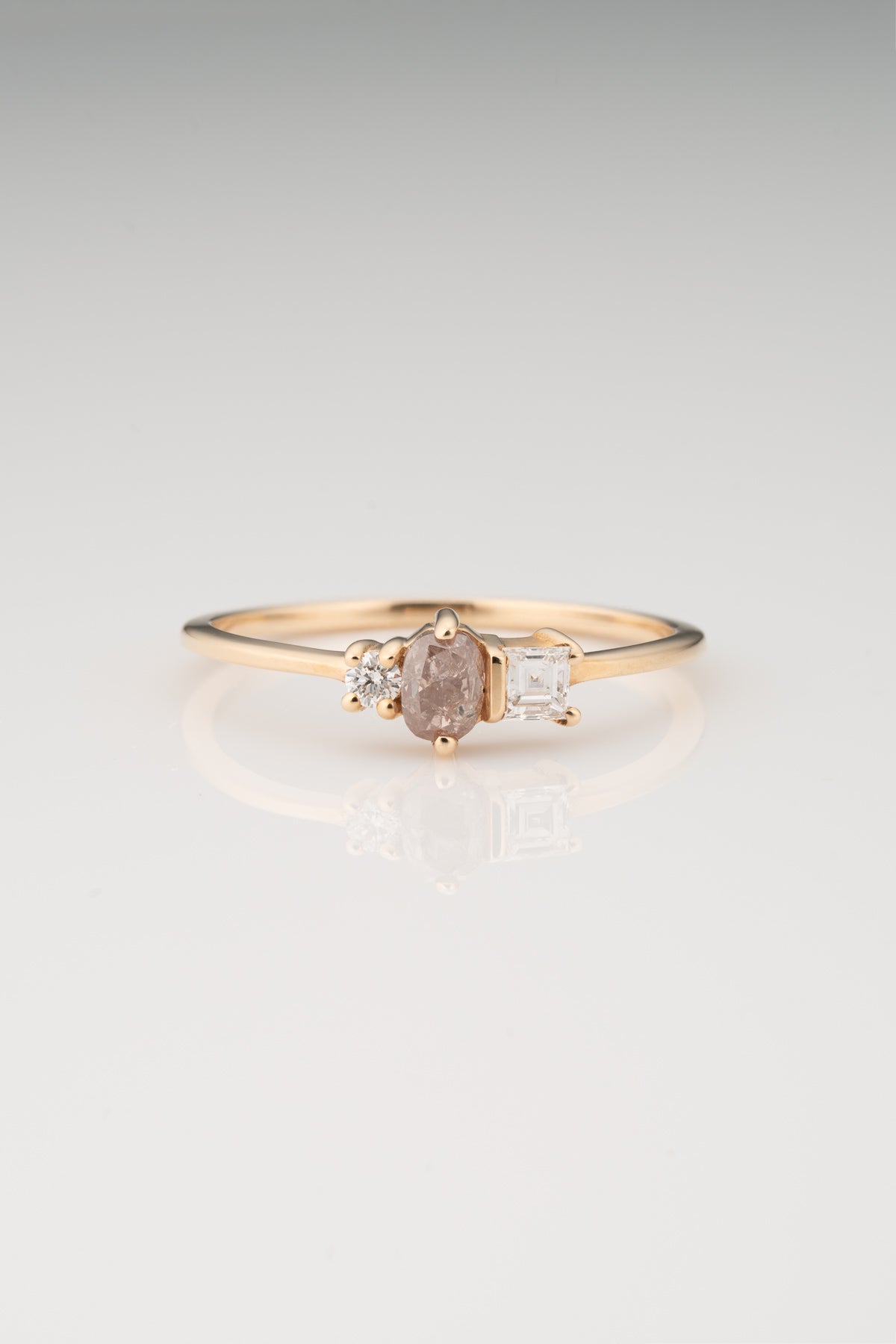 A Petite Gold Ring featuring a Oval Pink Diamond, a Round White Diamond, and a Square Asscher Cut Diamond in a mixed prong and bar setting on a white background. 