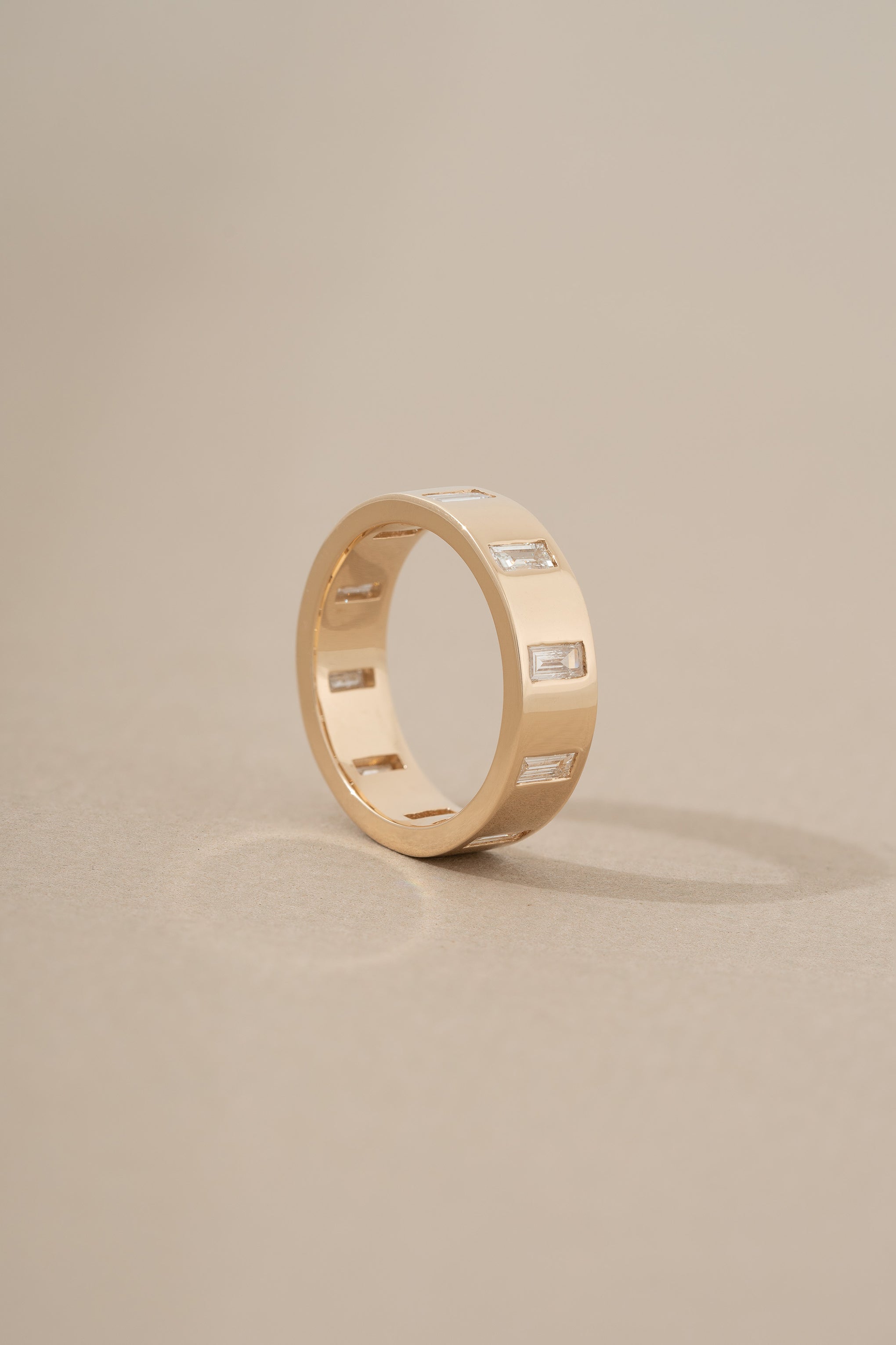 A thick gold band ring with north-south facing baguette cut diamonds gypsy set around its circumference on a neutral background